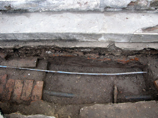 Foto 9: Escalón enterrado bajo el asfalto de la calle, cimiento de mampostería y la tierra original del terreno en la parte inferior. Nótese el contrapiso saliente original.
